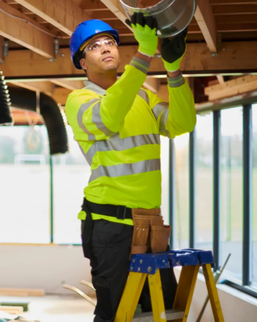 Un técnico con casco y chaleco reflectante, subido a una escalera, monta un conducto de ventilación en el techo de una obra