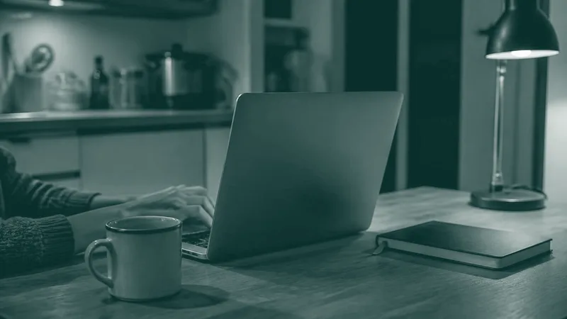 Un portátil abierto sobre una mesa de noche, con una lámpara encendida y una taza al lado