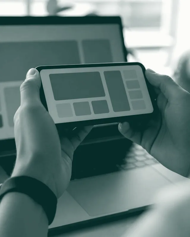 Unas manos sostienen un móvil con la interfaz de una app delante de un portátil